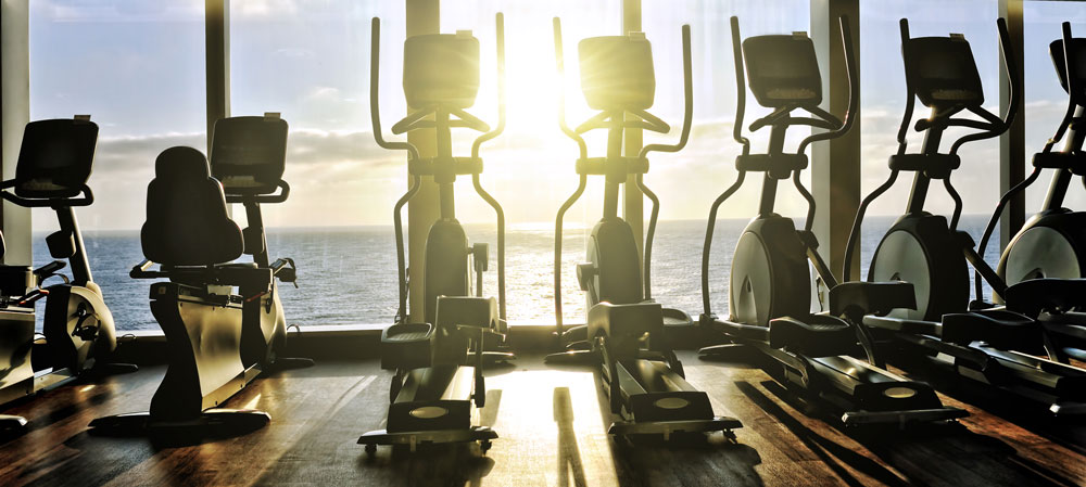 Intérieur de salle de fitness sombre avec des rangées d'appareils elliptiques et de vélos face à de grandes fenêtres donnant sur l'océan, ambiance bleu marine apaisante